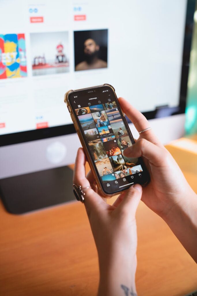 Close-up of a person browsing a smartphone with an Instagram feed, with a computer monitor in the background.